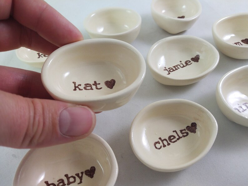 May include: A set of small, white ceramic bowls with brown lettering and hearts. The bowls are labelled with the names "kat", "jamie", "chelse", "baby", and other names.