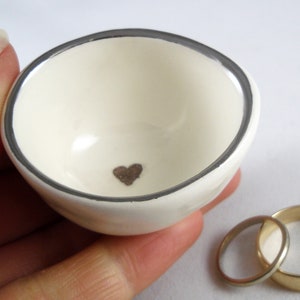 May include: White ceramic ring dish with a silver rim and a small brown heart in the center.