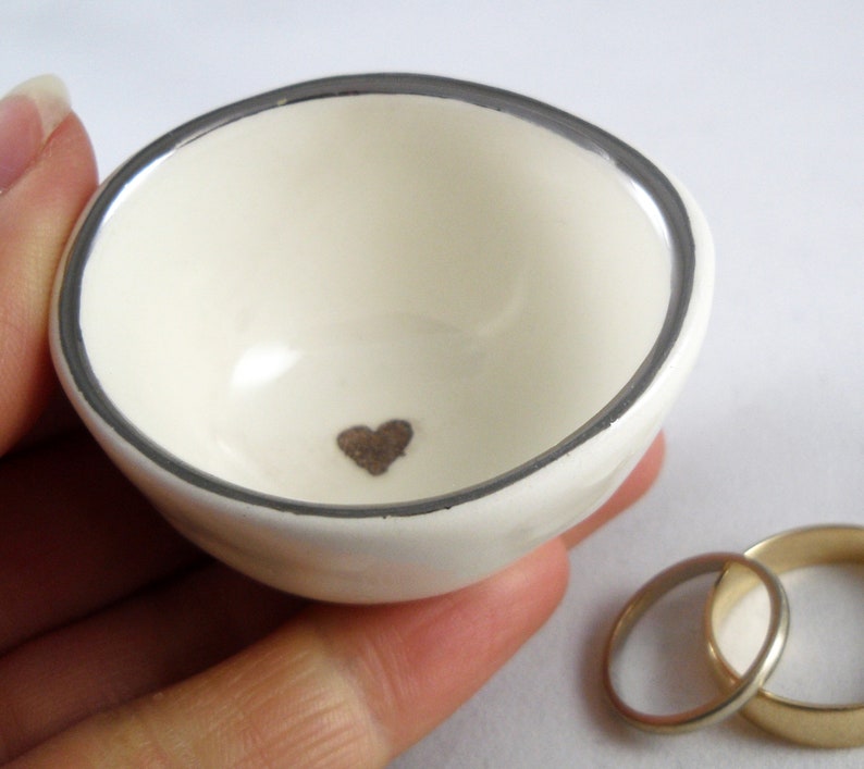 May include: White ceramic ring dish with a silver rim and a small brown heart in the center.