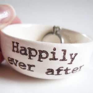 May include: White ceramic ring dish with the words "Happily ever after" printed in brown. A silver ring is sitting in the dish.