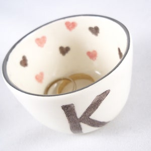 May include: White ceramic ring dish with a black letter K and pink and brown hearts. Two gold rings are inside the dish.