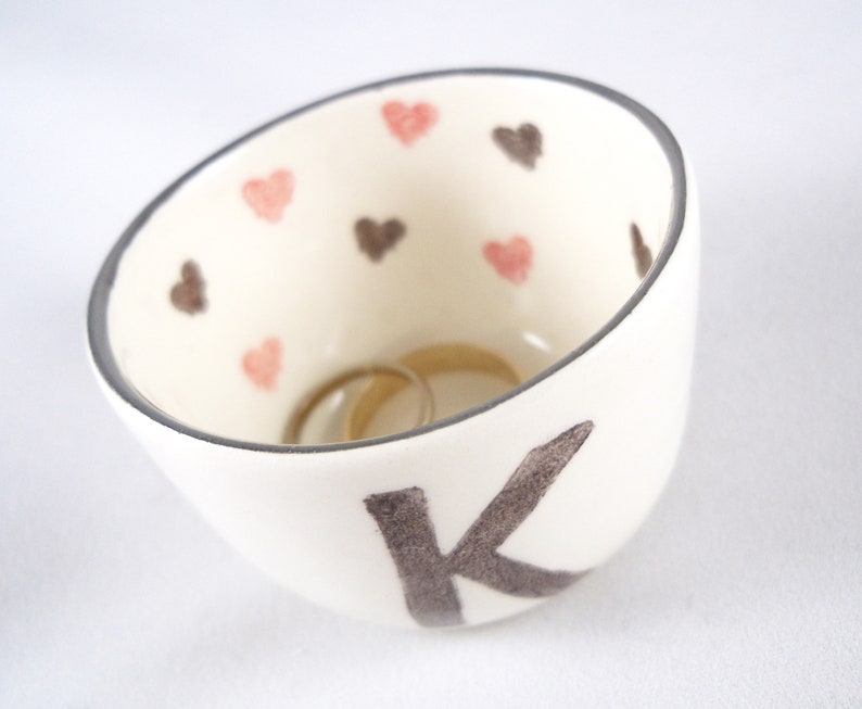 May include: White ceramic ring dish with a black letter K and pink and brown hearts. Two gold rings are inside the dish.