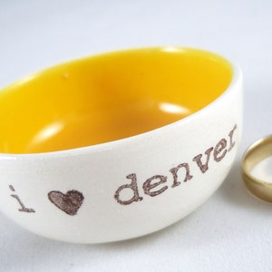 Puede incluir: Un pequeño tazón de cerámica blanca con un interior amarillo. El tazón tiene las palabras "i love denver" escritas en el lateral en letras marrones. Hay un símbolo de corazón entre las palabras "i" y "love". Dos anillos de oro están sentados junto al tazón.