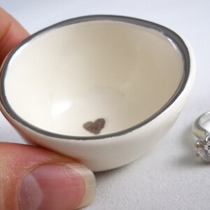 May include: A white ceramic ring dish with a silver rim and a small heart design in the center. The dish is sitting on a white surface.