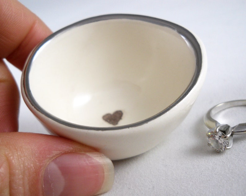 May include: A white ceramic ring dish with a silver rim and a small heart design in the center. The dish is sitting on a white surface.