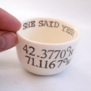 May include: Cream-colored ceramic bowl with the text "SHE SAID YES!" and coordinates 42.3770&deg;N 71.1167&deg;W in black. The bowl is held by a hand, showcasing its rounded shape and interior.