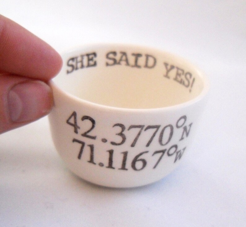 May include: Cream-colored ceramic bowl with the text "SHE SAID YES!" and coordinates 42.3770&deg;N 71.1167&deg;W in black. The bowl is held by a hand, showcasing its rounded shape and interior.