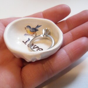 May include: White ceramic ring dish with a blue bird and the word "love" painted on it. A silver ring is resting in the dish.