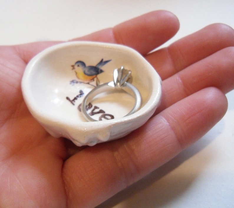 May include: White ceramic ring dish with a blue bird and the word "love" painted on it. A silver ring is resting in the dish.