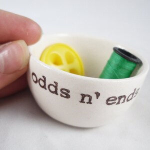 May include: White ceramic bowl with the words "odds n' ends" printed on the side. The bowl contains a yellow button and a spool of green thread.