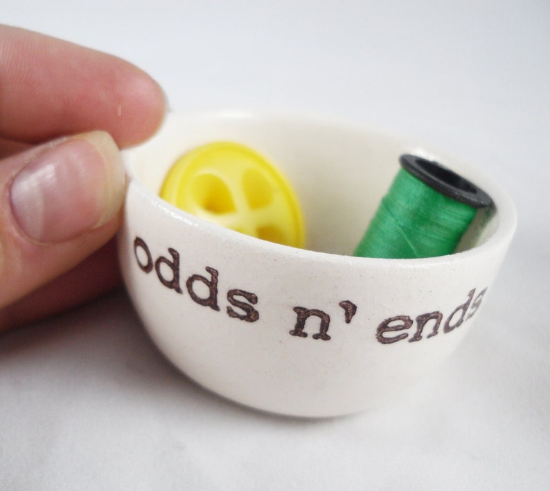 May include: White ceramic bowl with the words "odds n' ends" printed on the side. The bowl contains a yellow button and a spool of green thread.