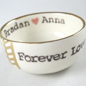 May include: A white ceramic bowl with a gold rim and a black inscription that reads "Bradan Anna Forever Love" with a small pink heart between the names.