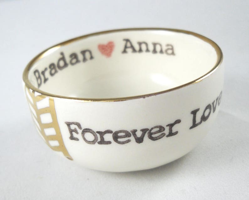 May include: A white ceramic bowl with a gold rim and a black inscription that reads "Bradan Anna Forever Love" with a small pink heart between the names.