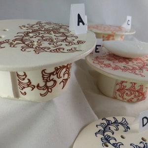 May include: Four ceramic soap dishes with different colored designs. The dishes are labeled A, B, C, and D. The dish labeled A has a brown design, the dish labeled B has a red design, the dish labeled C has a blue design, and the dish labeled D has a yellow design.