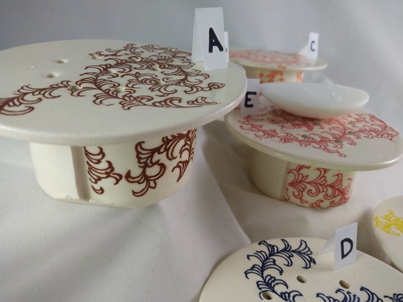 May include: Four ceramic soap dishes with different colored designs. The dishes are labeled A, B, C, and D. The dish labeled A has a brown design, the dish labeled B has a red design, the dish labeled C has a blue design, and the dish labeled D has a yellow design.