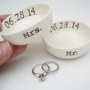 Puede incluir: Dos platos de anillos de cerámica blanca con las palabras "Mr." y "Mrs." y la fecha "06.28.14" inscritas en ellos. Un par de alianzas de plata están sobre una superficie blanca.