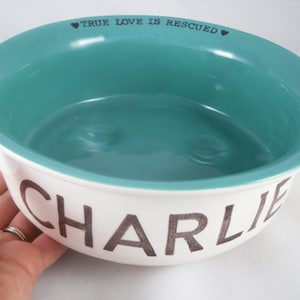 May include: White ceramic dog bowl with a teal blue interior. The bowl has the text "TRUE LOVE IS RESCUED" printed on the rim and the name "CHARLIE" printed on the bottom.