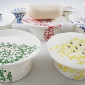 May include: A set of nine white ceramic soap dishes with various floral designs. Each dish has a raised base and drainage holes. The designs include green, yellow, blue, and red floral patterns.