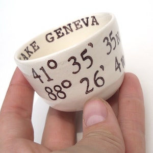 May include: A small, off-white ceramic bowl with the words "LAKE GENEVA" and coordinates 41&deg; 35' N, 88&deg; 26' 4W written in brown ink. The bowl has a slightly angled shape and a smooth surface.