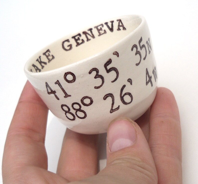May include: A small, off-white ceramic bowl with the words "LAKE GENEVA" and coordinates 41&deg; 35' N, 88&deg; 26' 4W written in brown ink. The bowl has a slightly angled shape and a smooth surface.