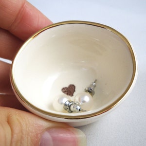 May include: A small white ceramic dish with a gold rim, featuring a heart design and two pearl earrings.