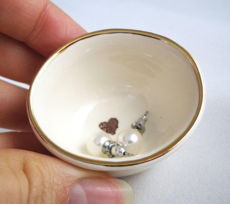 May include: A small white ceramic dish with a gold rim, featuring a heart design and two pearl earrings.