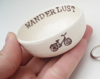 May include: A small, cream-coloured ceramic bowl with the word "WANDERLUST" printed in dark brown. A bicycle illustration is also printed on the side. The bowl is held in a hand, with a silver ring visible in the background.