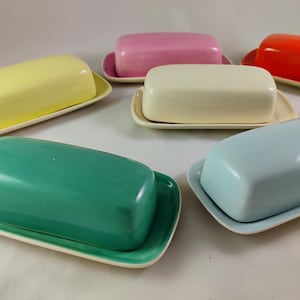 以下が含まれることがあります： 黄色、ピンク、オレンジ、白、青、緑など、さまざまな色合いのカラフルなセラミックバター皿のコレクション。各皿には長方形の蓋と、それに合わせた長方形のベースが付いています。バター皿は白い表面に配置されています。