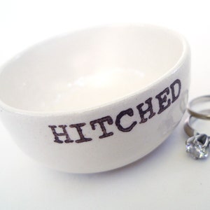 May include: White ceramic ring dish with the word "HITCHED" printed in black. Two silver rings are sitting in the dish.