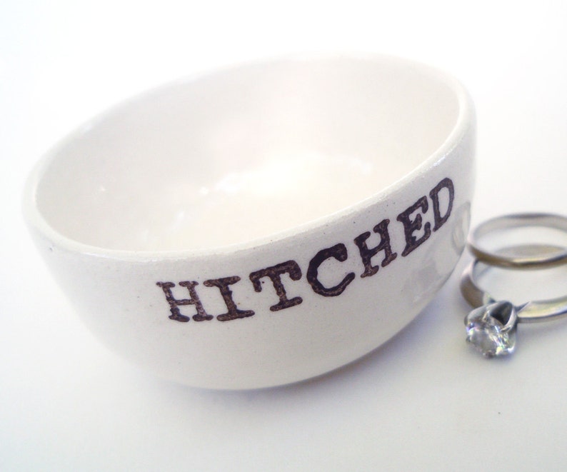 May include: White ceramic ring dish with the word "HITCHED" printed in black. Two silver rings are sitting in the dish.