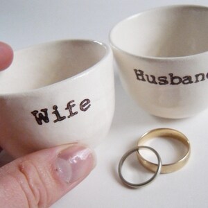 Puede incluir: Dos cuencos de cer&aacute;mica blanca con las palabras "Wife" y "Husband" escritas en ellos. Un anillo de boda de oro y plata est&aacute; en primer plano.