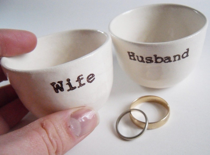 Puede incluir: Dos cuencos de cer&aacute;mica blanca con las palabras "Wife" y "Husband" escritas en ellos. Un anillo de boda de oro y plata est&aacute; en primer plano.