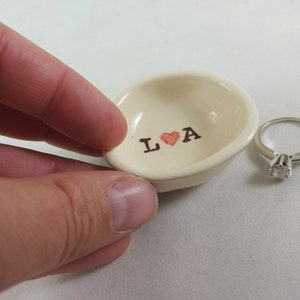 May include: A white ceramic ring dish with the words "L &hearts; A" printed in brown. A silver ring with a clear stone is sitting next to the dish.