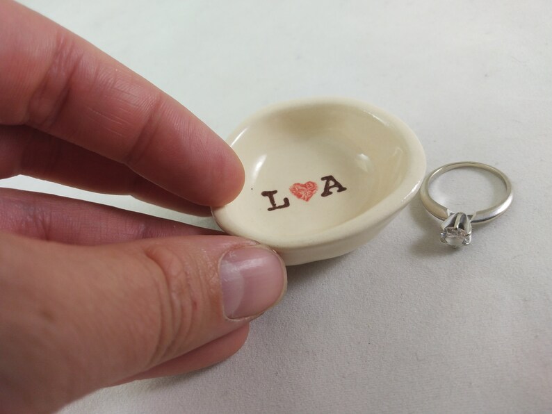 May include: A white ceramic ring dish with the words "L &hearts; A" printed in brown. A silver ring with a clear stone is sitting next to the dish.