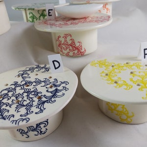 May include: Five ceramic soap dishes with different colored floral designs. The dishes are labeled with letters D, E, and F.