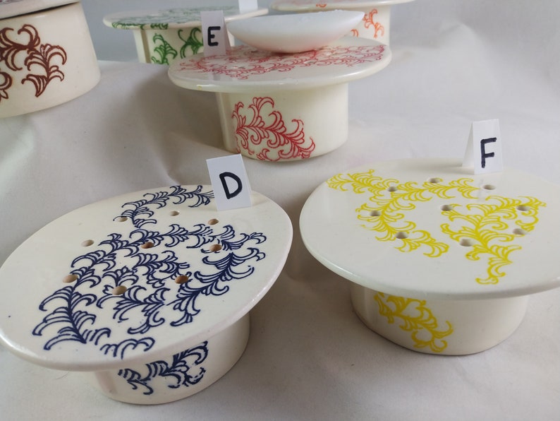 May include: Five ceramic soap dishes with different colored floral designs. The dishes are labeled with letters D, E, and F.