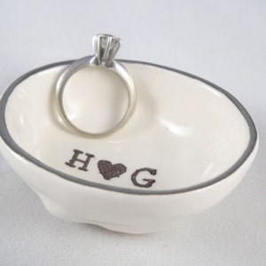 May include: White ceramic ring dish with a silver rim. The dish has the words "H &hearts; G" inscribed on the bottom. A silver ring is sitting on the dish.