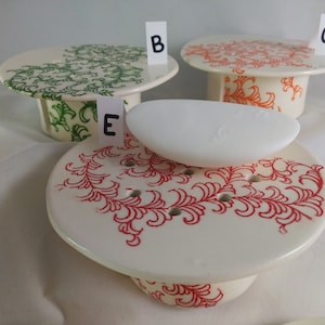 May include: Four ceramic soap dishes with different designs. The dishes are white with red, green, and orange floral patterns. One dish has a white bar of soap on top.
