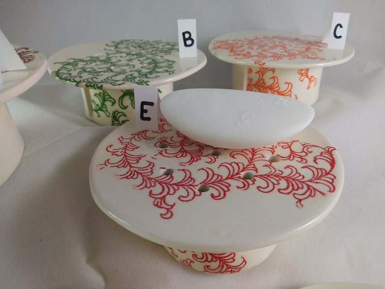 May include: Four ceramic soap dishes with different designs. The dishes are white with red, green, and orange floral patterns. One dish has a white bar of soap on top.