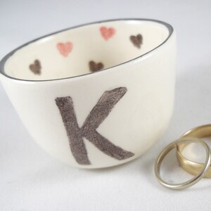 May include: White ceramic ring dish with a gray letter K and pink and brown hearts around the rim. Two gold wedding bands are sitting on a white surface next to the dish.