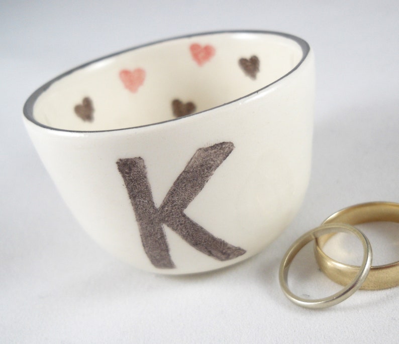 May include: White ceramic ring dish with a gray letter K and pink and brown hearts around the rim. Two gold wedding bands are sitting on a white surface next to the dish.