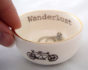May include: White ceramic ring dish with a gold rim. The dish has the word "wanderlust" written on the inside and a black and white illustration of a tandem bicycle on the outside.