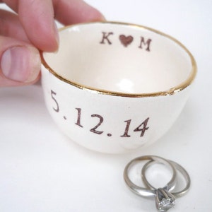 Puede incluir: Plato de cerámica blanco para anillos con borde dorado. El plato está inscrito con las iniciales "K ♥ M" y la fecha "5.12.14".