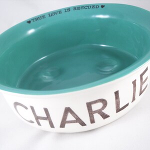 May include: White ceramic pet bowl with a teal blue interior. The bowl has the words "TRUE LOVE IS RESCUED" printed on the rim and the name "CHARLIE" printed on the bottom in a silver-colored font.