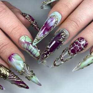May include: A set of long, pointed artificial nails with intricate designs. The nails feature a combination of colours, including purple, green, and gold. Some nails have a marbled effect, while others have gold filigree accents.