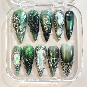 May include: A set of ten artificial nails in a clear plastic case. The nails feature a variety of designs, including swirling green and black patterns, silver accents, and iridescent elements. The nails are in various shapes.