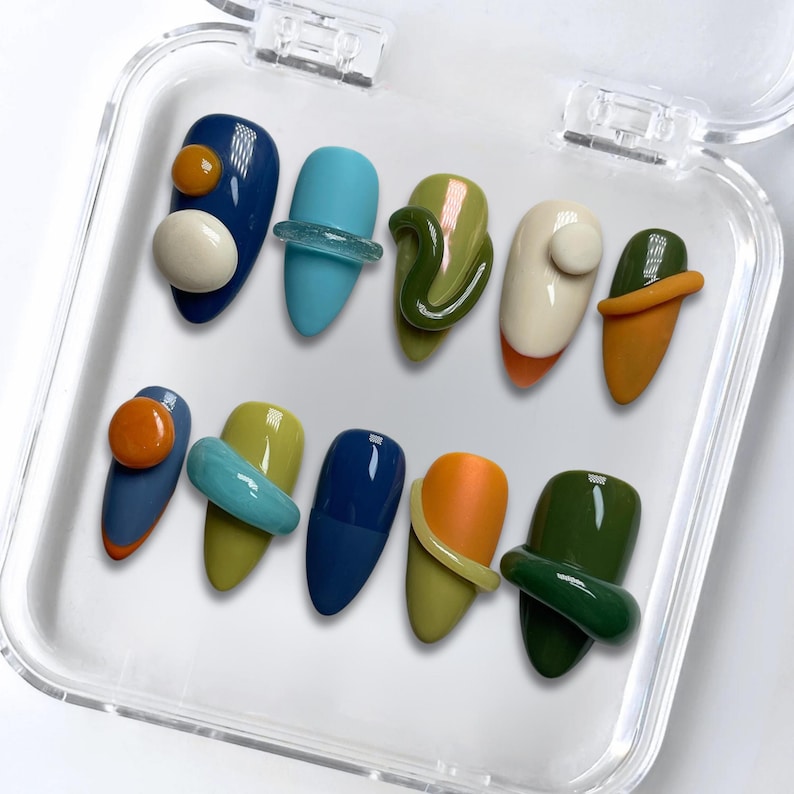 May include: A set of ten artificial nails in a clear plastic case. The nails are painted in various colors, including blue, green, orange, and white, with abstract 3D designs. The nails are almond-shaped.