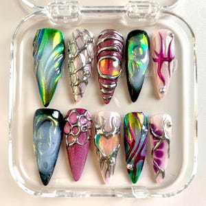 May include: A set of ten long, pointed artificial nails in a clear plastic case. Each nail features a unique design with iridescent colors, metallic accents, and intricate patterns. The color palette includes green, pink, silver, and black.