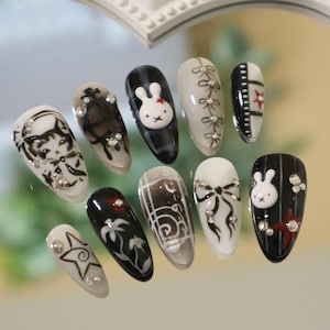 Puede incluir: Una colección de diez uñas artificiales con varios diseños. Las uñas presentan bases negras, blancas y transparentes con obras de arte detalladas, incluyendo un conejito, lazos y patrones florales. Algunas uñas están adornadas con pedrería.
