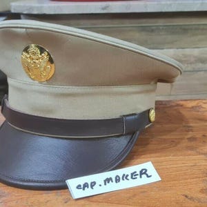 Puede incluir: Un sombrero de estilo militar beige con una visera y una banda marrón oscuro. El sombrero presenta un emblema dorado y una pequeña etiqueta que dice "CAP. MAKER". El sombrero está sobre una superficie de madera.